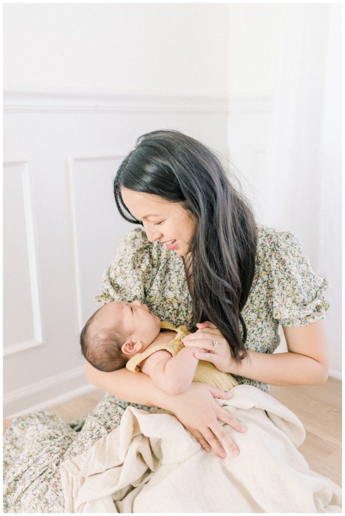 A photo of a young mom wearing a ditsy olive green floral dress sitting on hardwood floors in a natural lit, white studio holding her one month old son looking down at him while holding his hand.