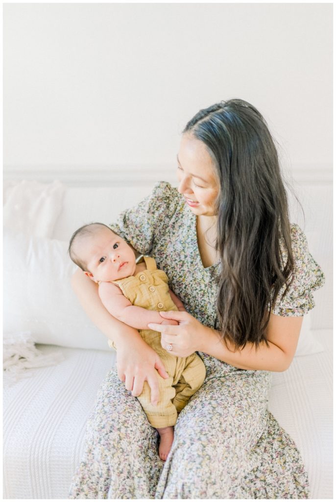 Photo of a young mom with long dark hair sitting on a white couch in a studio with natural light looking down at her newborn son wearing light yellow overalls holding his hand while he looks at the camera.