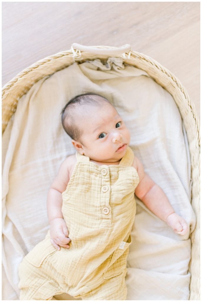 Photo of a newborn boy laying in a wicker basket on top of light hardwood floors looking up at the camera while wearing light yellow overalls.
