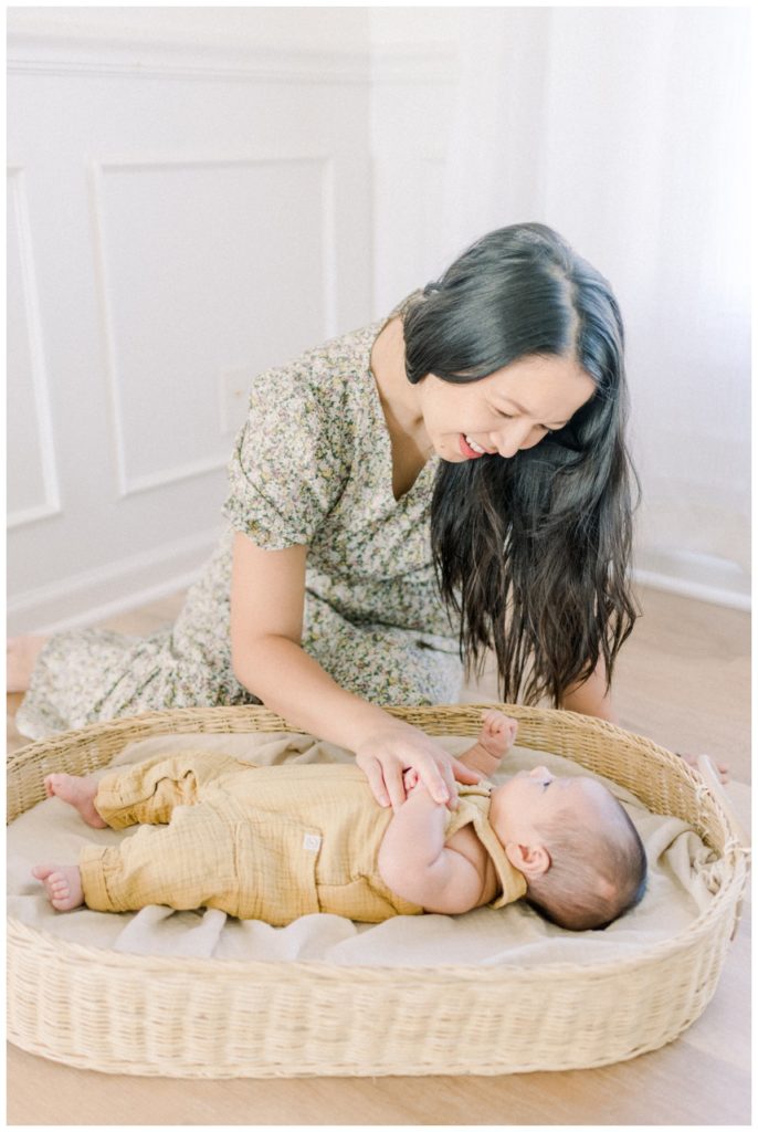 Photo of a young mom sitting on light hardwood floors in a natural light white studio looking down at her newborn son laying in a wicker basket holding his hand while he looks up at her wearing a cotton gauze light yellow romper.