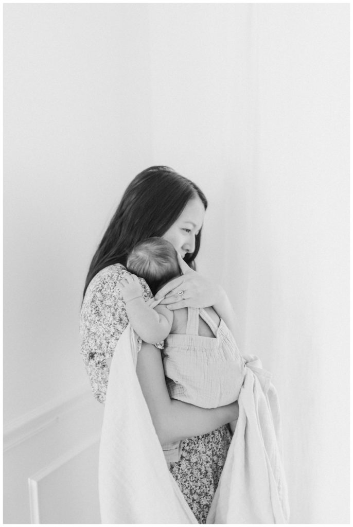 Black and white photo of a young mom standing up in a white studio by large windows holding her newborn son to her check soothing him with a light colored blanket draped around them.