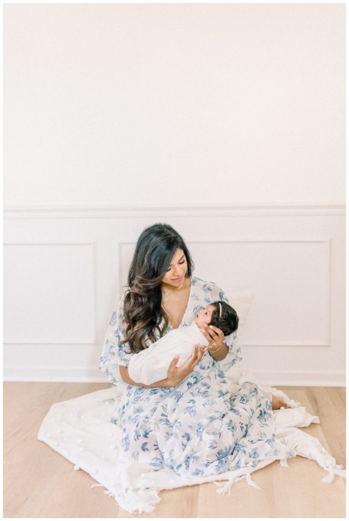 Image of young mom sitting on a well lit white newborn studio floor wearing a Baltic Born white and blue dress holding her two week old daughter.