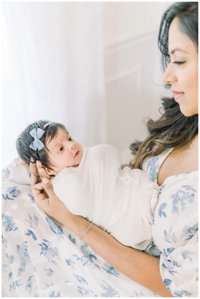 A photo of a young mom sitting by a soft lit window holding her newborn daughter swaddled in a white wrap with a small blue bow.