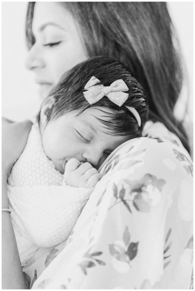 A black and white photo of a young mom soothing her newborn swaddled daughter against her shoulder.