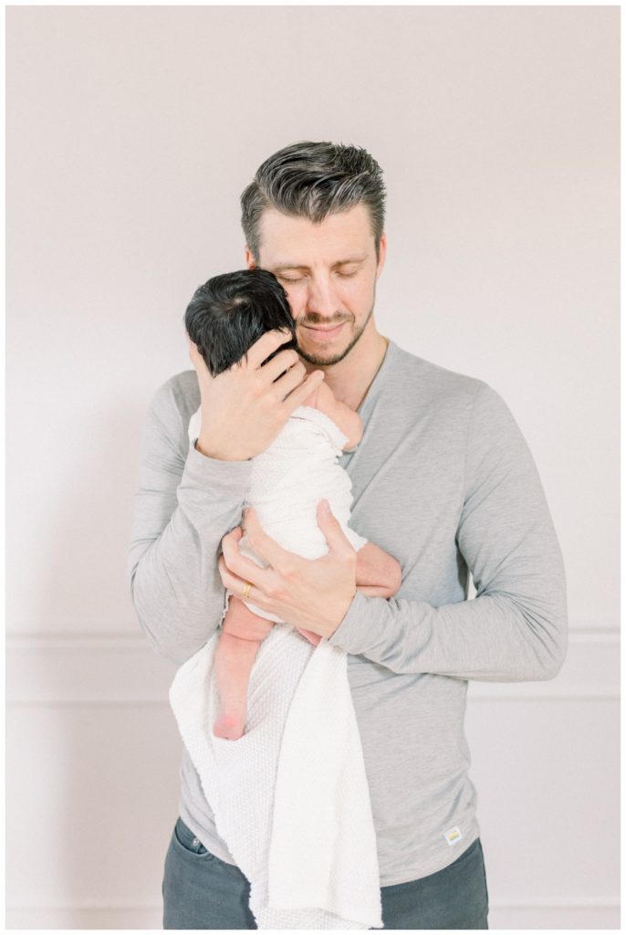 An image of a young dad standing in a white studio with natural light holding his newborn daughter check to cheek with eyes closed soothing her.