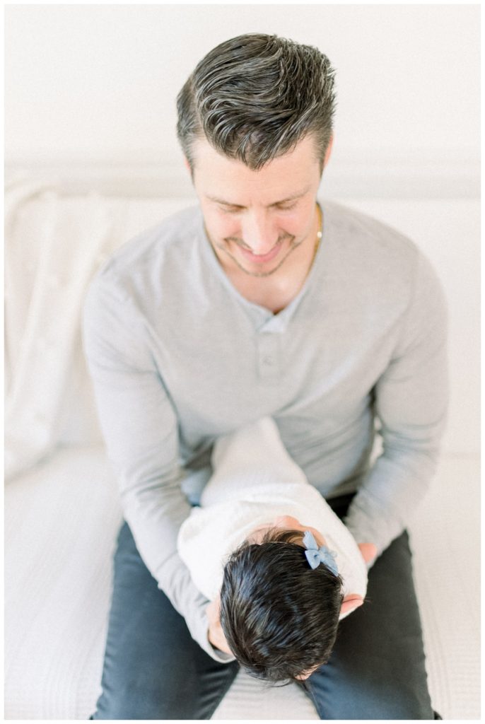 Photo of a young dad holding his newborn daughter facing him looking down at her in a white swaddle wrap and blue bow sitting on a white couch. 