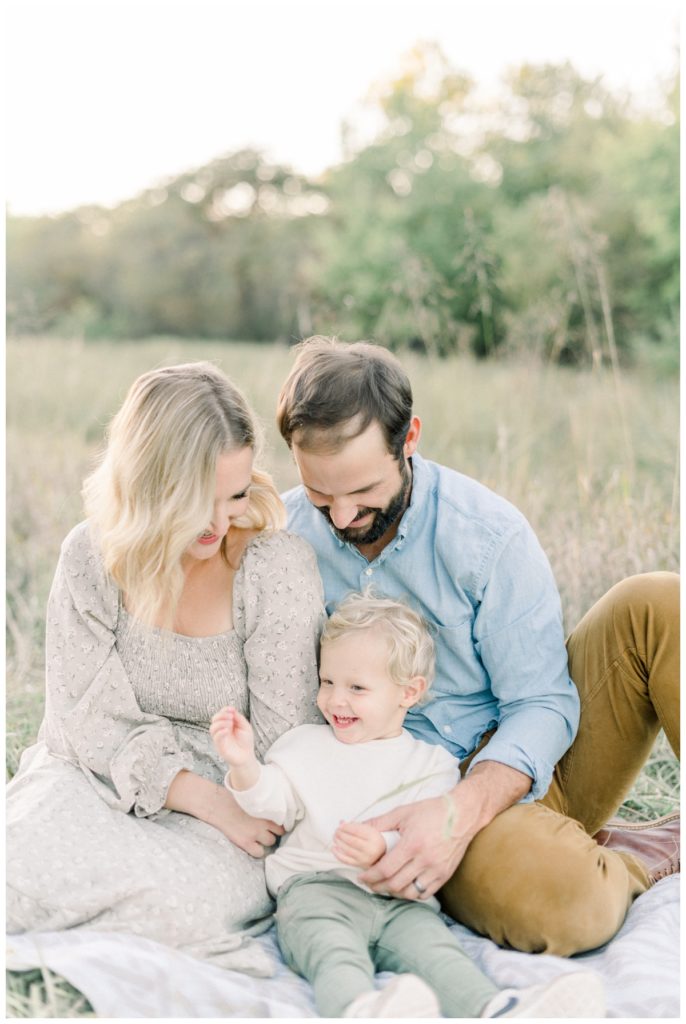 Photo of a family of three sitting in a field while the mother wears a soft green floral dress and toddler son smiles while wearing a cream sweater and olive pants.