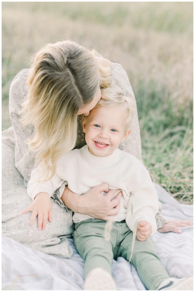 Photo of a mother and her toddler son who is wearing a cream sweater and olive pants smiling at the camera while his mother hugs him and kisses the top of his head. 