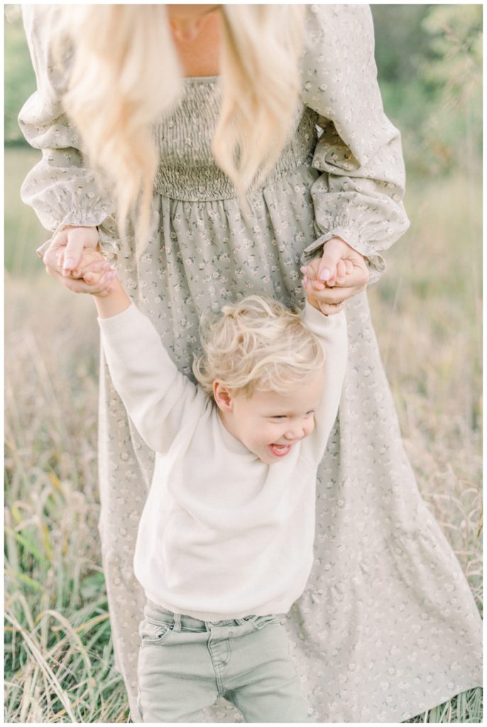 A playful photo of a mom wearing a floral green dress holding both the hands of her son at sunset in a field while he smiles wide. 