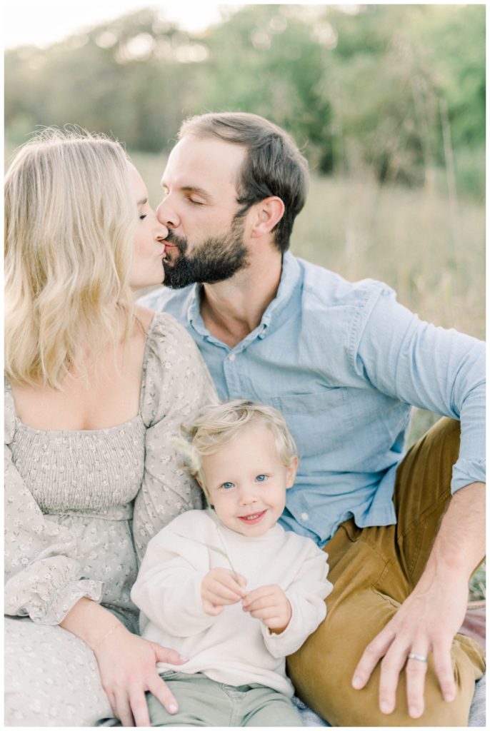 Photo of a family of three sitting in a field on a blanket while the parents kiss above their son's head and he is wearing a cream sweater looking directly at the camera. 