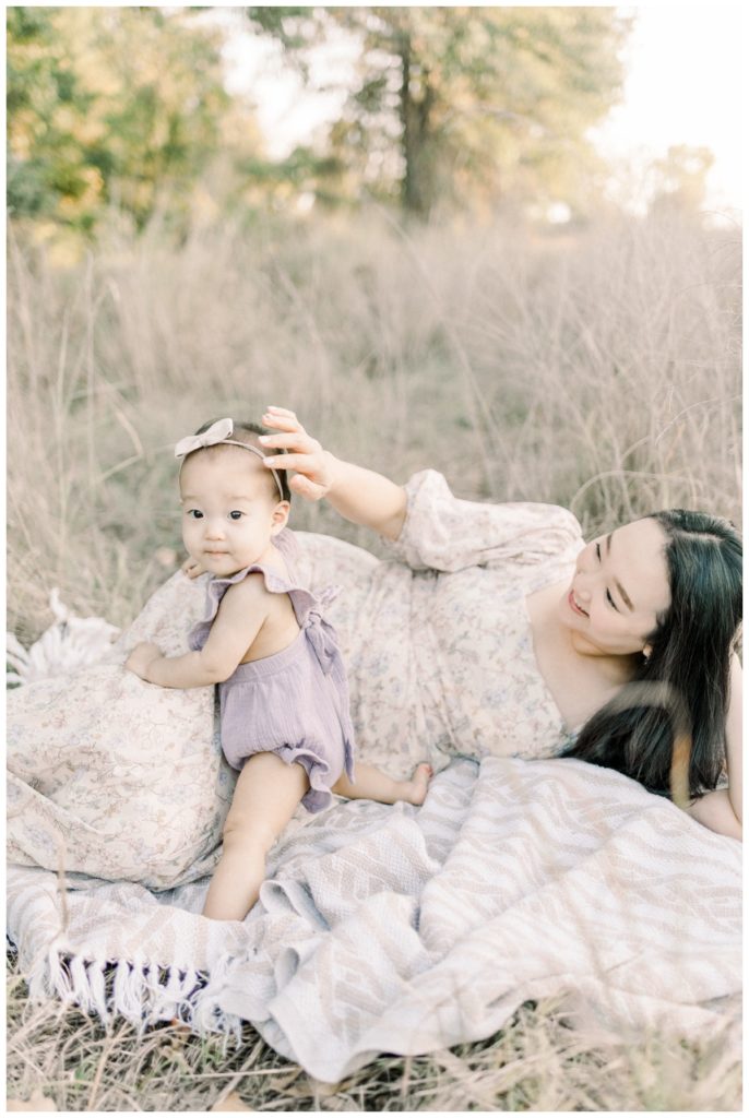 Photo of a young mom wearing a soft lavender and cream floral dress laying on a blanket in a wheat field with her newborn daughter beside her.