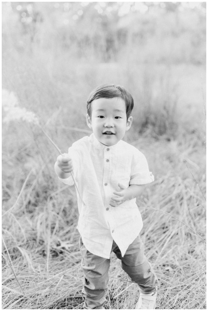 Black and white photo of a toddler boy looking at the camera while holding a piece of wheat grass. 