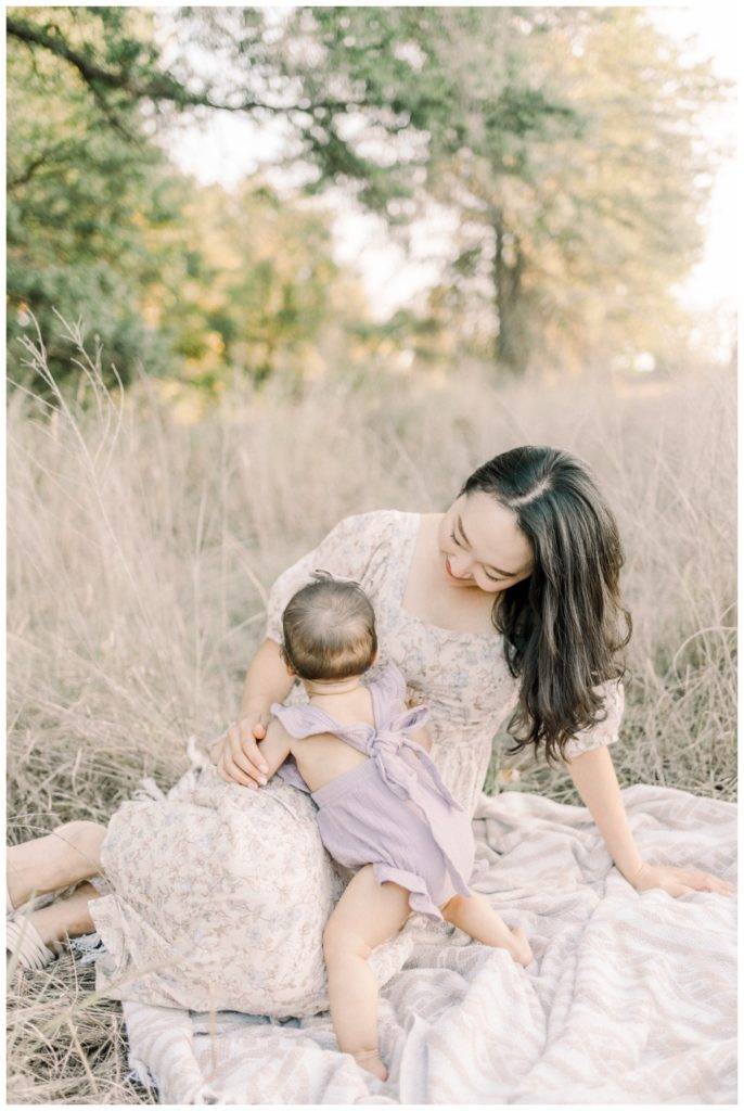 A photo of a young mom wearing a cream floral dress sitting on a blanket in a wheat field while looking lovingly down at her newborn daughter.