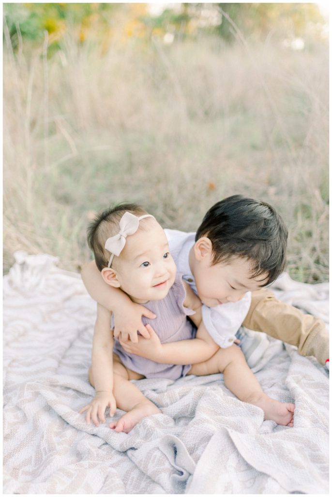 Photo of young siblings sitting in a wheat field on a blanket with the older brother wrapping his arms around his younger sister who is wearing lavender and a cream bow for a hug. 