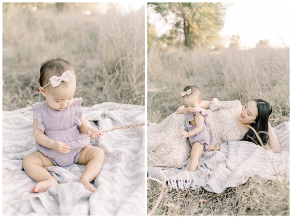 A close up photo of a six month old girl sitting on a blanket in a wheat field wearing a soft lavender romper and cream bow playing with a piece of grass.