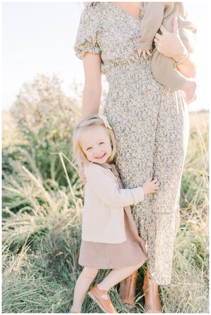 Photo of a toddler girl wearing a mauve dress and cream sweater hugging her mom's leg. 