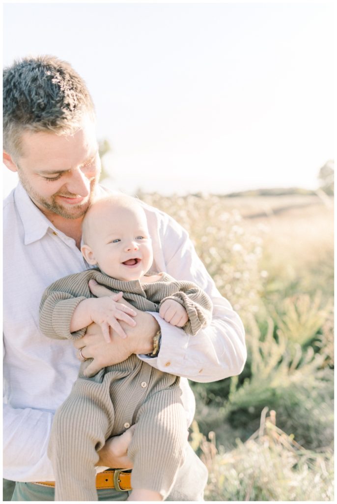 A picture of a young dad holding his six month old son to his check looking down at him in a field at sunset. 