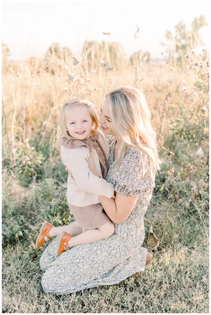 Picture of a young mom sitting on her knees in a field at sunset holding her daughter on her lap who is wearing a mauve dress and cream sweater.