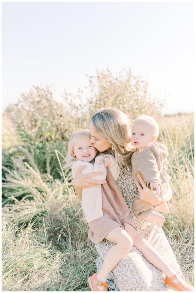 A young blonde mom holding both her toddler daughter in a mauve dress and cream sweater and six month old son in a brown romper sitting in a beautiful field kissing her daughter's head. 