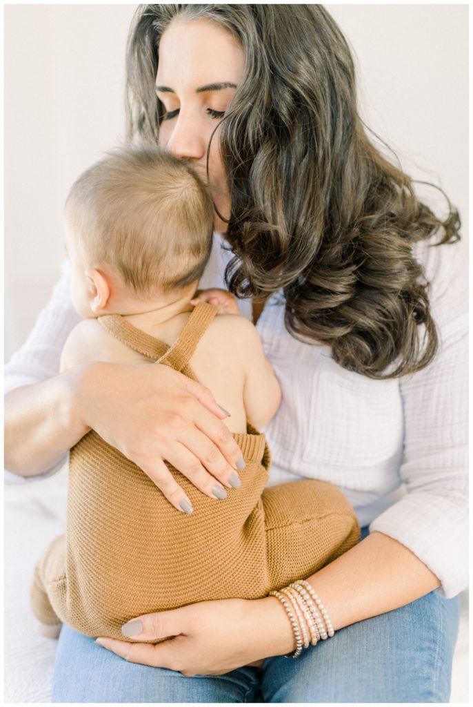 Photo of a young mom holding her nine month old son wearing a cotton copper criss cross romper chest to chest kissing the side of his head in a white natural lit room. 