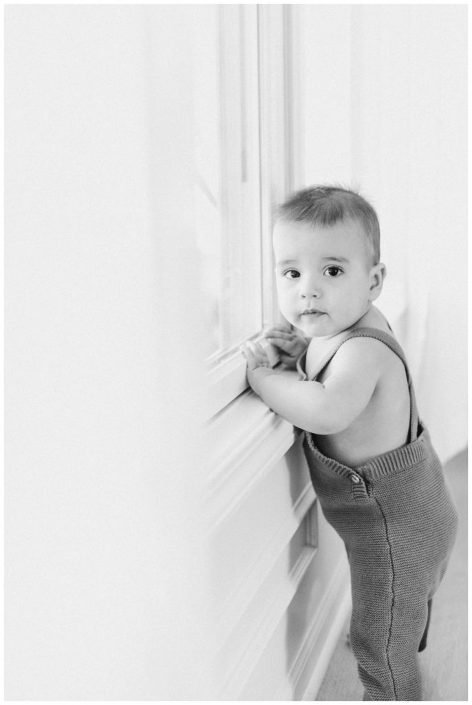 Black and white photo of a baby standing at the window wearing a romper beside white curtains looking at the camera. 