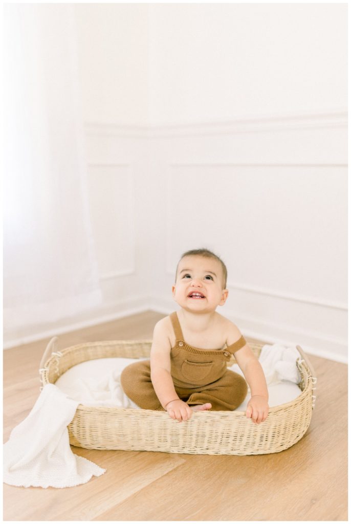 Picture of a natural light studio space with white walls and white curtains with a nine month old baby sitting in a wicker basket.