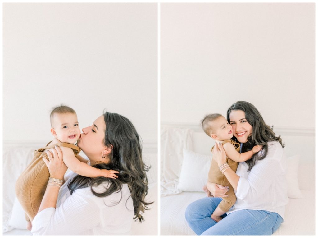 Picture of a young mom wearing a white button down shirt holding her son who is wearing a copper romper and kissing his cheek in a white studio. 
