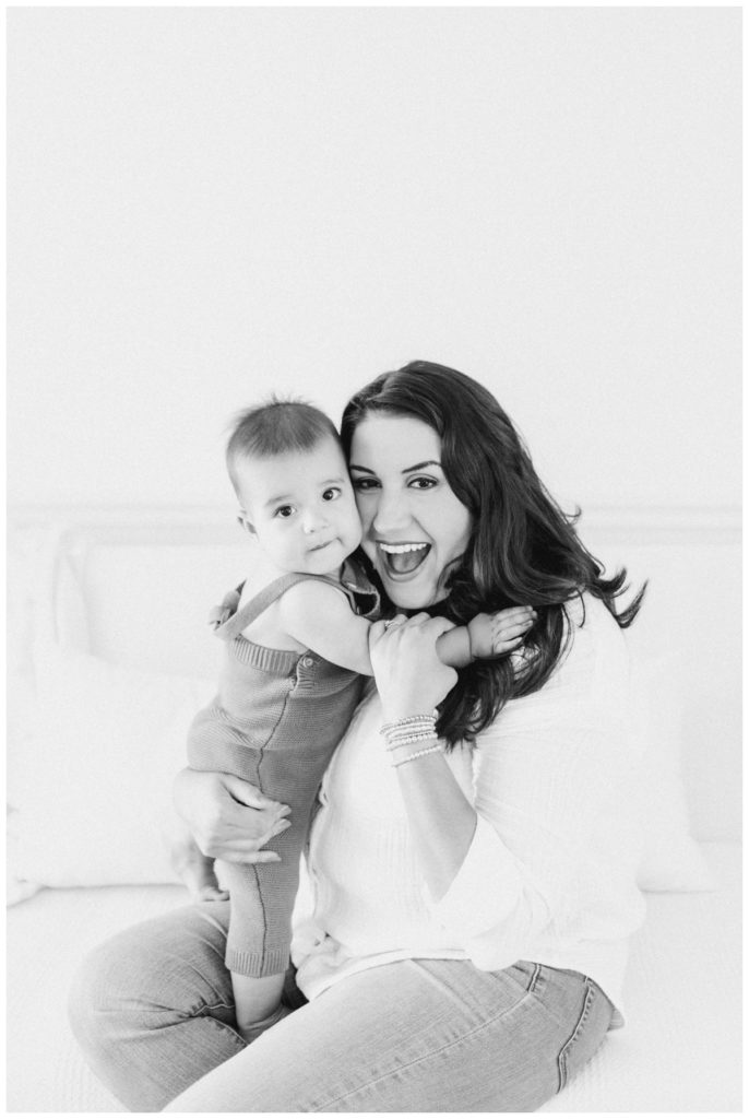 Black and white photo of a young mom wearing a white button down shirt and light wash jeans holding her nine month old baby close to her chest while they are both smiling and looking at the camera. 