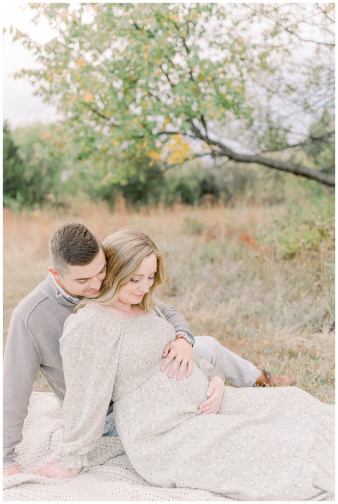 Picture of a young couple sitting on a blanket in a wheat field with Fall toned colors of trees with the wife leaning against the husband while both of them have their hand on her expectant belly. 
