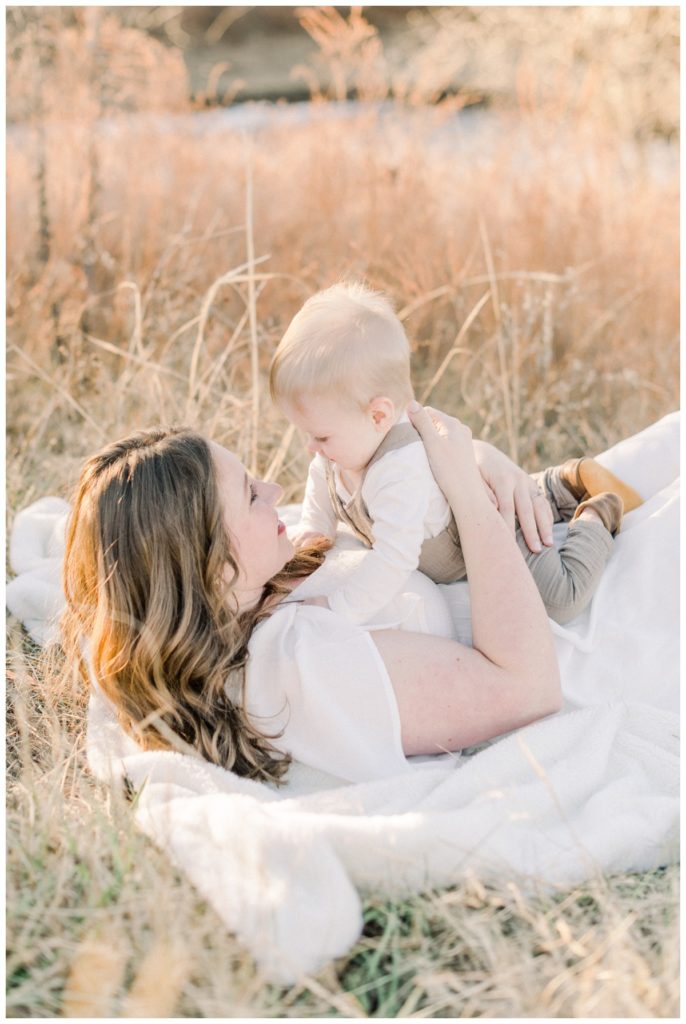 Picture of a mom in a white dress laying on white blanket in the field while her six month old son lays on her stomach and looks down at her. 
