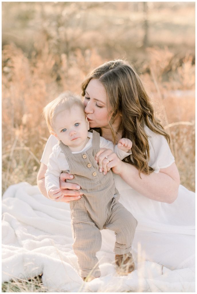 Picture of a mom wearing a flowy white dress sitting on a cream blanket in a golden field kissing the side of her young son's head while he stands beside her. 