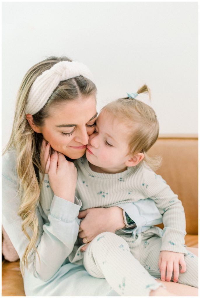 Photo of a young mom wearing a cream headband holding her two year old daughter on her lap while sitting on a cognac leather couch snuggling cheek to cheek. 
