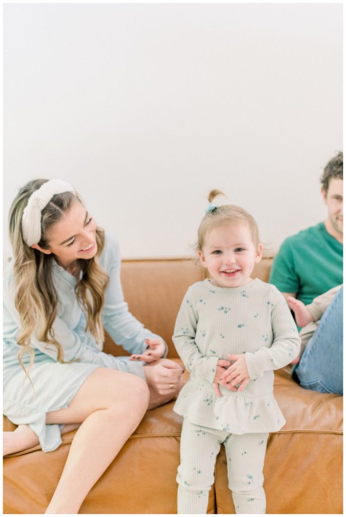 Picture of a young family sitting in their living room on a cognac leather couch playing playfully with their young daughter while she laughs. 