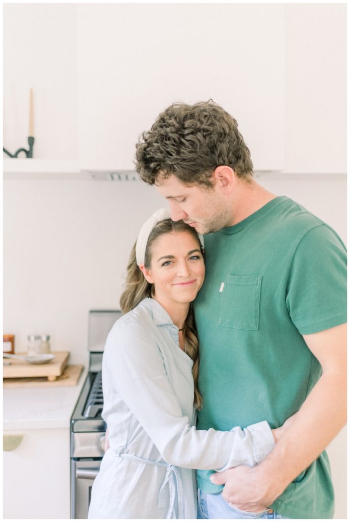 Photo of a young couple standing in a clean, white kitchen while she hugs him and wears a light jean dress and he looks down at her. 