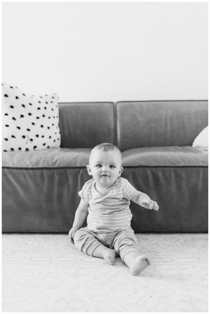 Black and white photo of a toddler boy sitting on the floor against the couch looking at the camera while wearing a striped shirt.