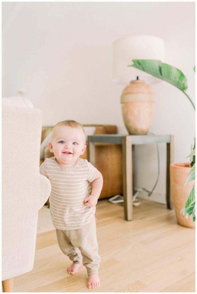 Photo of a toddler boy standing in a modern living room with a large floor plant to the side while he wears a striped cream and white shirt and leans against the couch. 