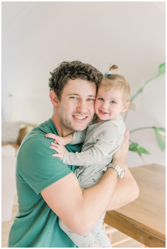 Photo of a dad wearing a sage green shirt holding his toddler daughter with blonde hair with a small ponytail as they sit in their white kitchen smiling and looking at the camera. 