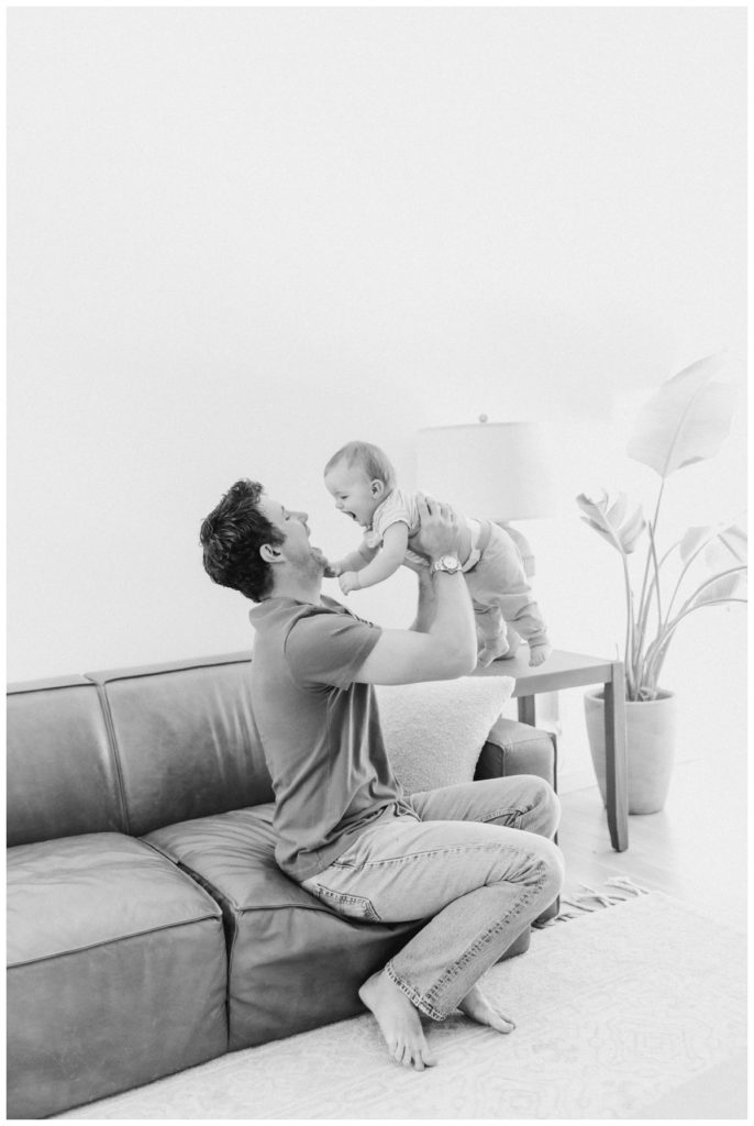 Black and white photo of a young dad sitting on the couch holding his toddler son up in the air. 