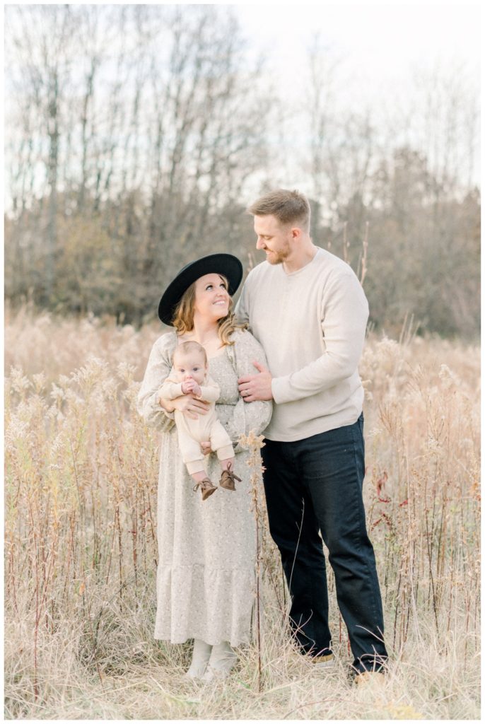 Photo of a family of three standing in a fall wheat field looking at one another while she wears a soft olive maxi dress and black hat, he is a in a cream sweater and black slacks and the baby is in an all cream onesie. 