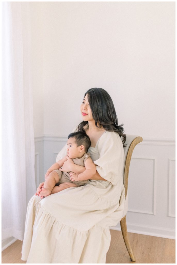 Picture of a young mom wearing a baby doll style cream dress sitting on a cream chair holding her six month old son with dark hair in a white studio looking out the window with natural light. 