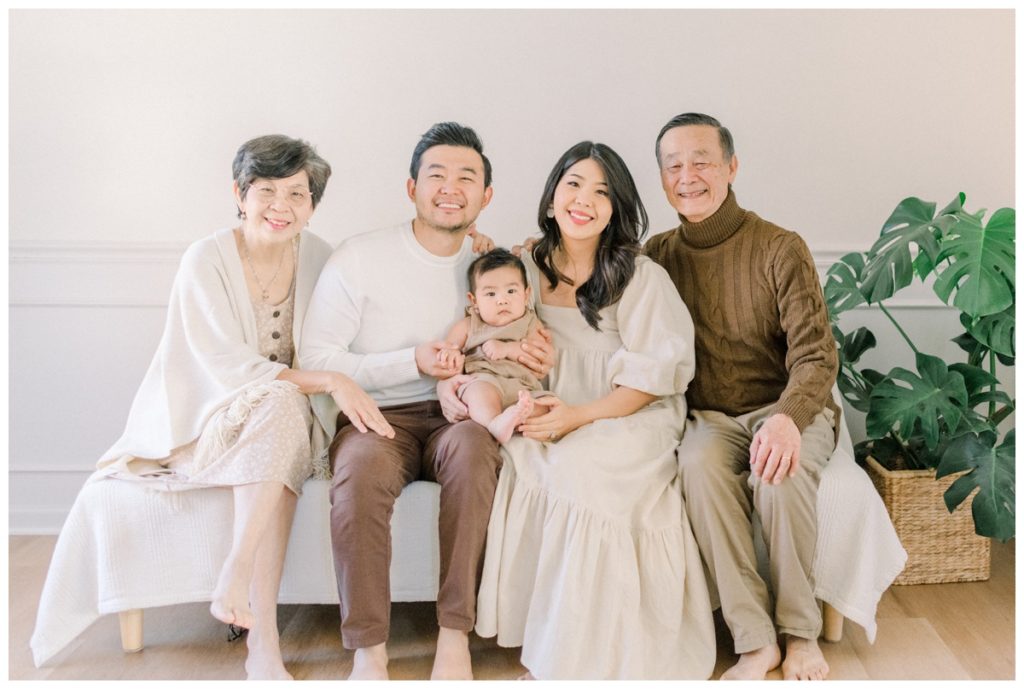 Group photo of an extended family sitting in a natural lit white studio room with wood floors all looking at the camera wearing different natural toned clothing. 