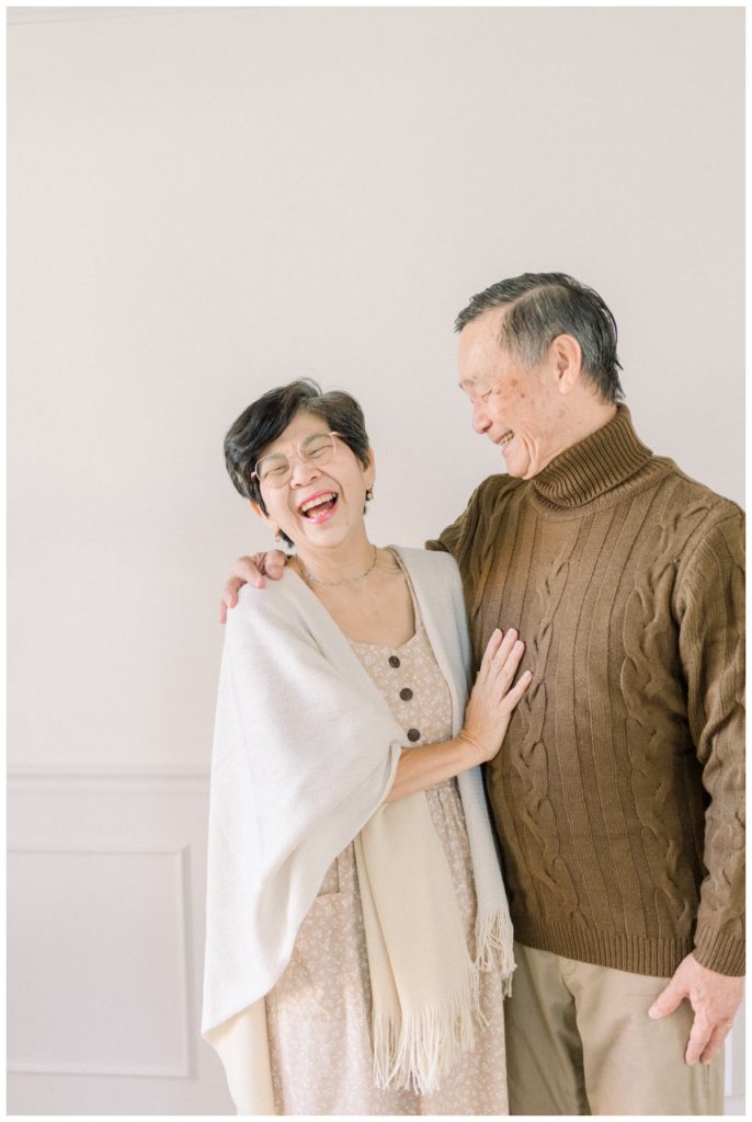 Photo of a middle aged couple where the husband has his arm around his wife's should looking at her while she has her hand on his chest laughing while they stand in a white studio space. 