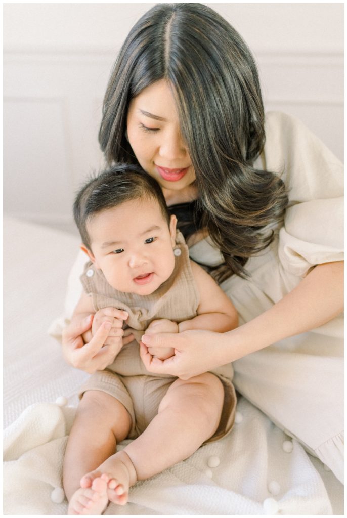 Close up photo of a young mom laying on her side holding the hands of her six month old son who is sitting on a cream dotted blanket in a white studio space. 