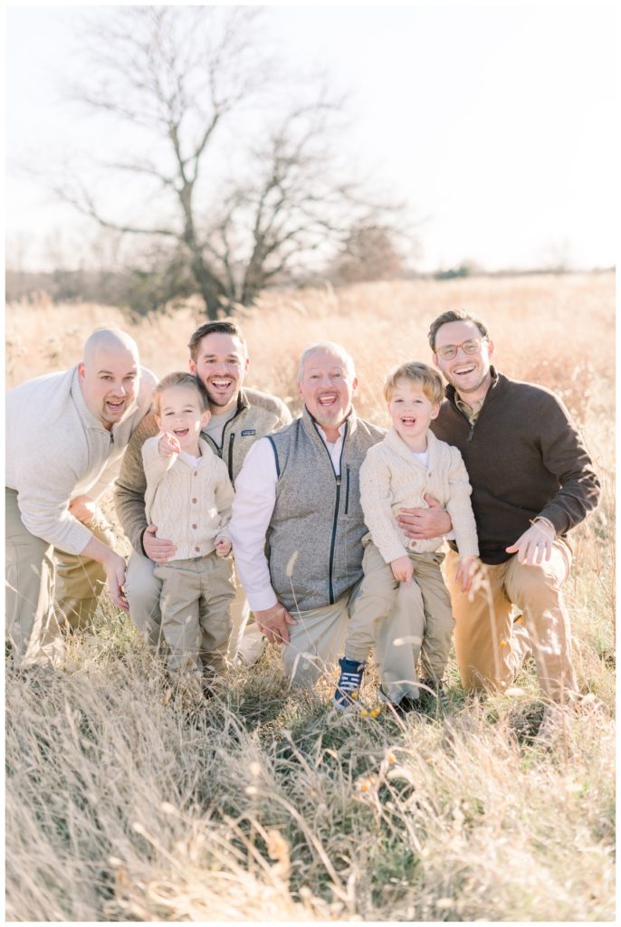Picture of three generations of men all kneeling down in a wheat field smiling and looking at the camera with various tones of neutral outfits. 