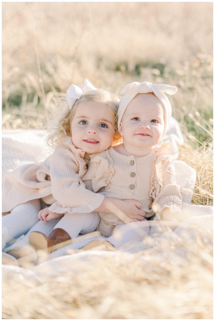 A close up picture of two little sisters sitting on a white blanket in a wheat field with the older sister's arm around the baby both wearing different neutral toned dresses. 
