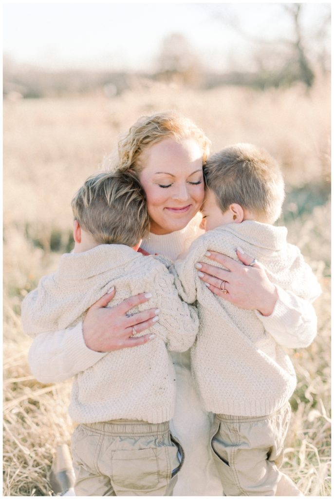 Photo of a young mom with blonde hair kneeling down in a wheat field closing her eyes while her two toddler sons both hug her at the same time. 