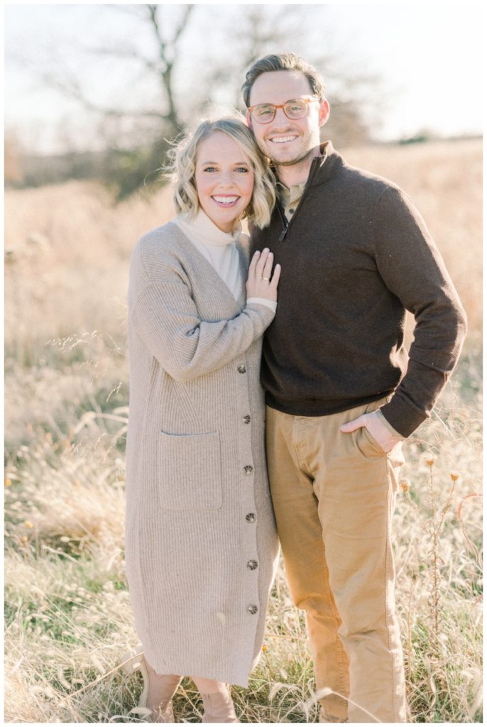 Picture of a young couple standing in a wheat field at golden hour side by side wearing neutral toned outfits smiling and looking at the camera. 