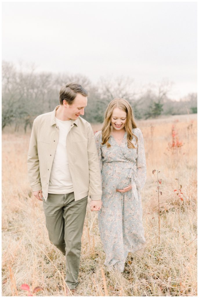 Picture of a young couple holding hands walking through a wheat field looking down at her stomach while she has one hand on it as they smile expecting their first baby. 