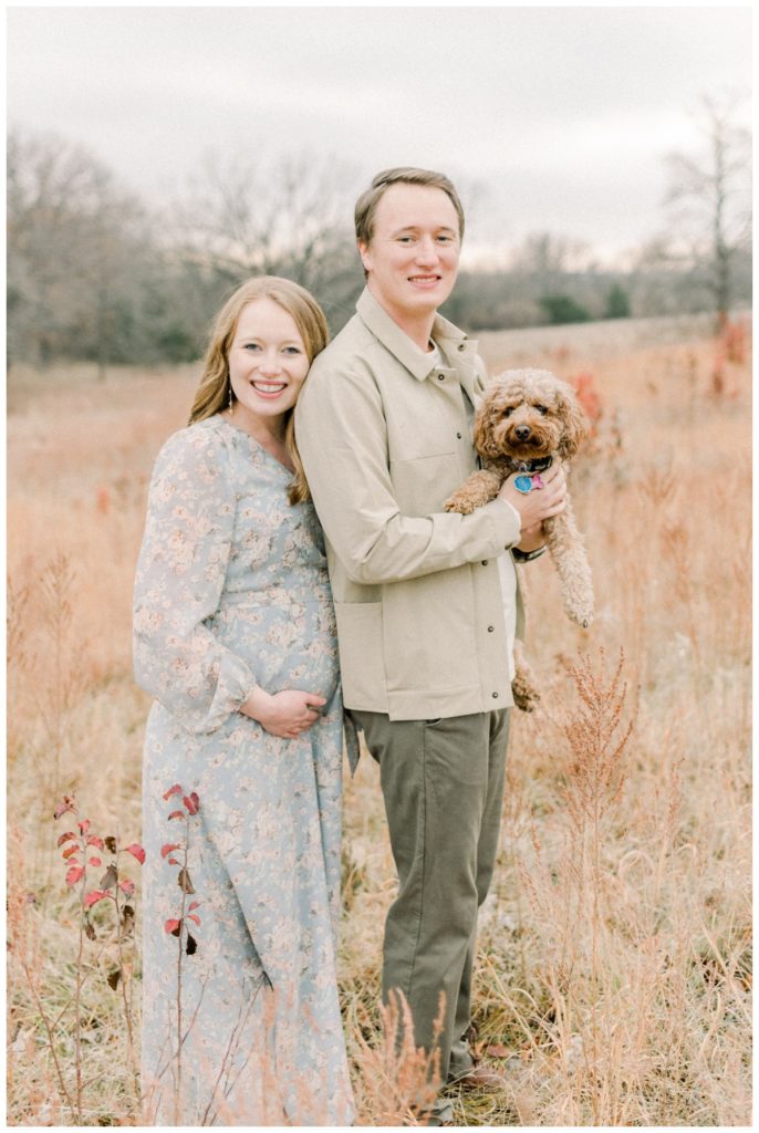 Photo of a young couple standing in a wheat field while the husband holds their small Golden Doodle puppy and the wife leans against his back with her head on his shoulder and hand on her expectant belly while they all look at the camera. 