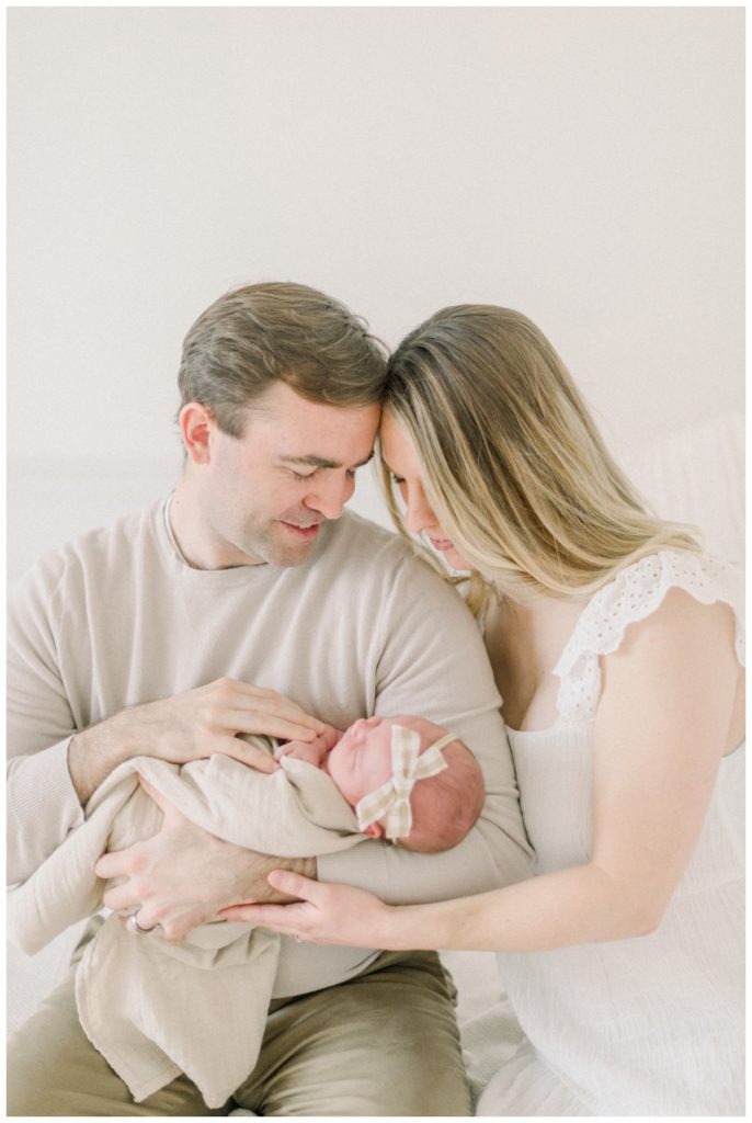 Mom and dad in a white natural light studio holding newborn baby in a cream swaddle with a plaid bow.